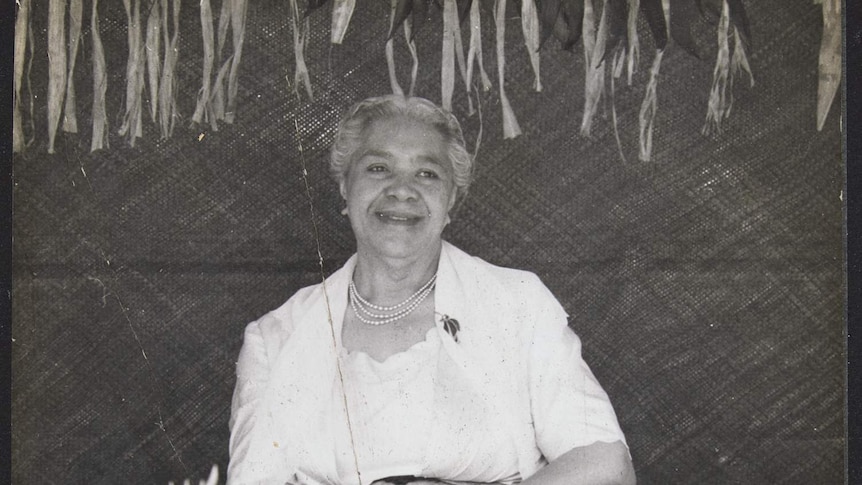 Queen Sālote sits on a chair smiling, presumably to an audience. Decorations of coconut palm branches surround.