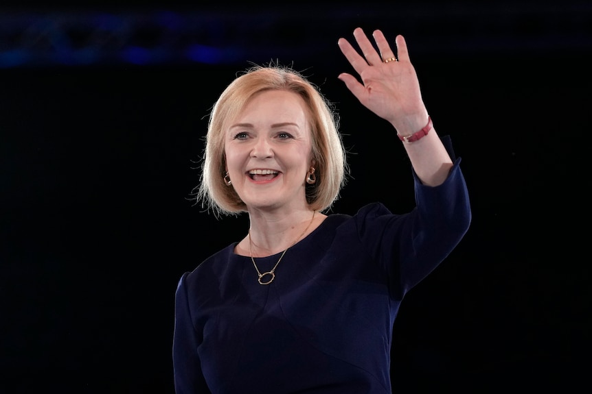 Liz Truss, in a navy blue dress, smiles and waves against a dark background.  She wears a red band wristwatch