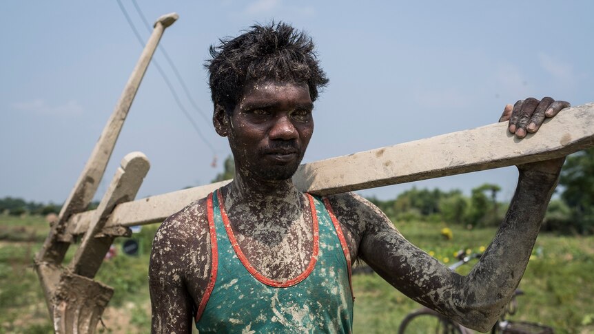 A farmer covered in mud carries a wooded plough on his shoulder.