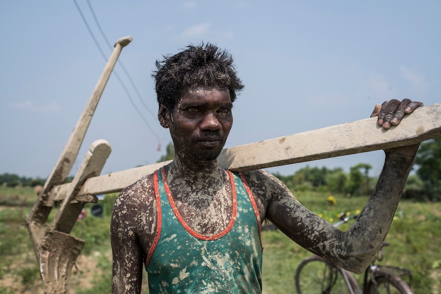 A farmer covered in mud carries a wooded plough on his shoulder.