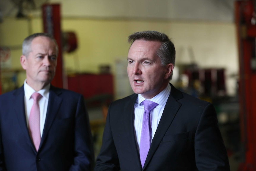 Chris Bowen, wearing a lavender tie, speaks while Bill Shorten watches in the background.