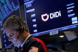 A stock trader with a headset looks down into a bright screen as a monitor behind him displays the "D" logo of company DiDi.