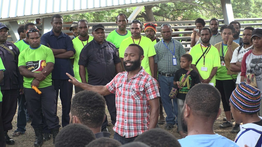 A man wears a red and white plaid shirt as he speaks to a group of men at a polling station in port moresby