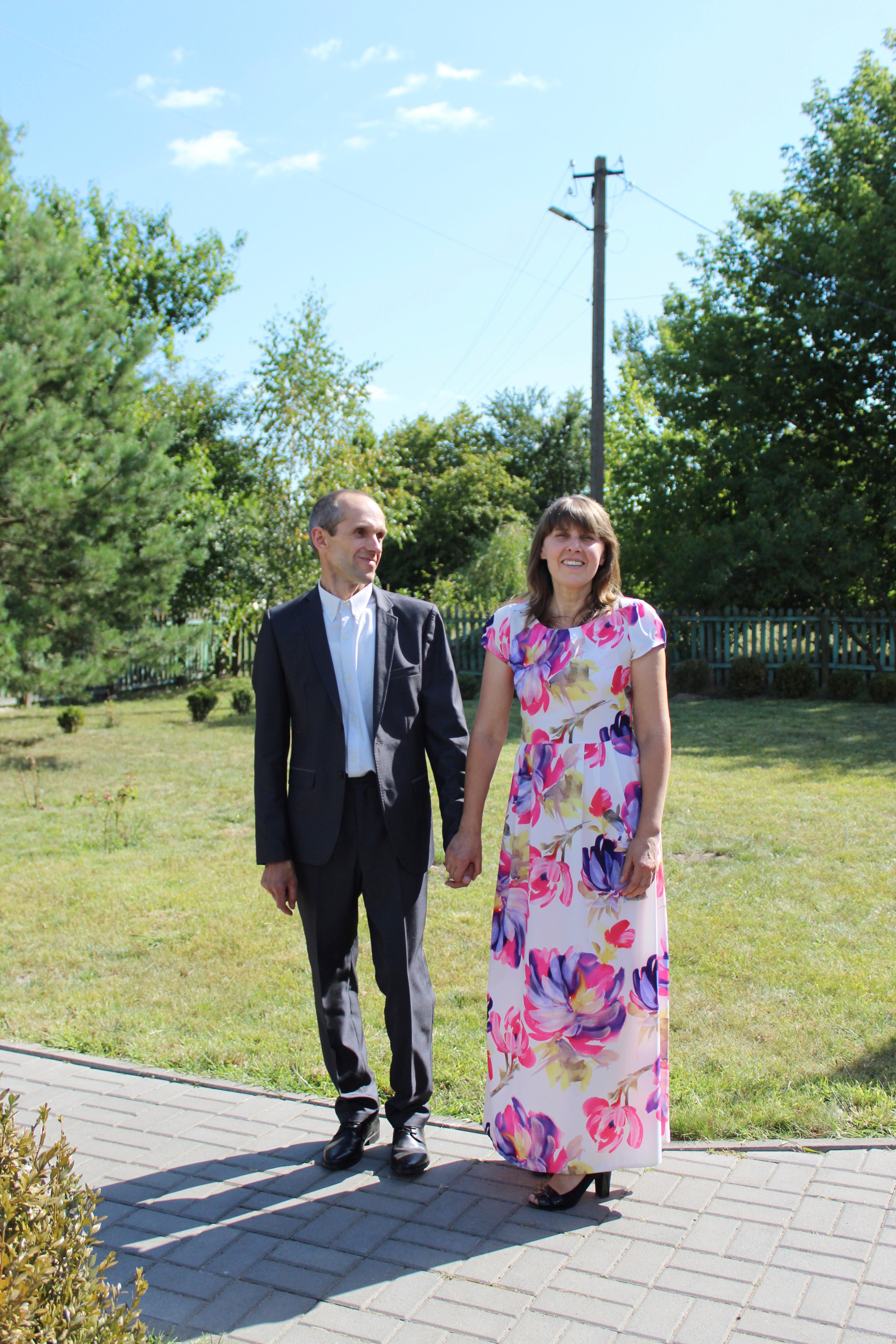 A man in a suit and a woman in a floral dress clasp hands, smiling in the sun. The man is looking at the woman 