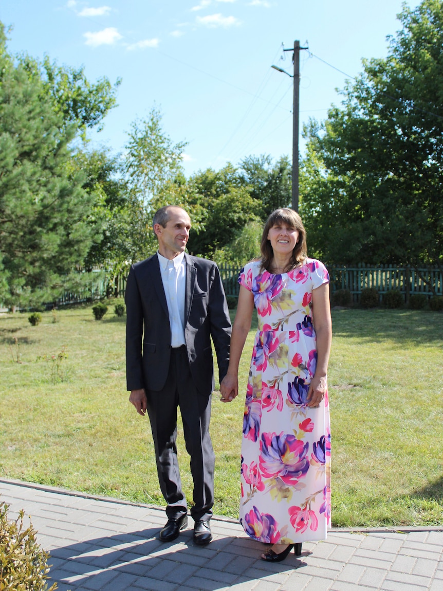 A man in a suit and a woman in a floral dress clasp hands, smiling in the sun. The man is looking at the woman 