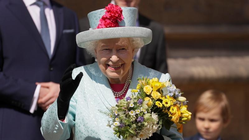 The Queen, wearing a blue suit-hat outfit, holds bouquets of flowers in one hand and waves with the other