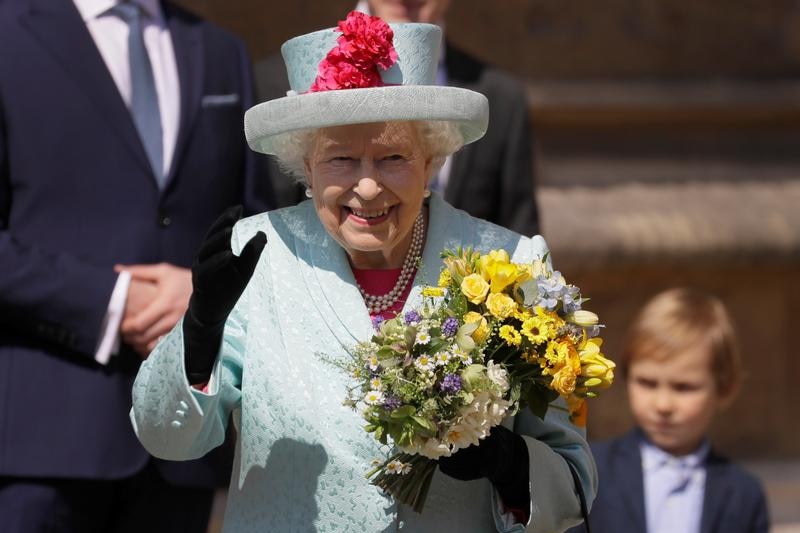 The Queen, wearing a blue suit-hat outfit, holds bouquets of flowers in one hand and waves with the other