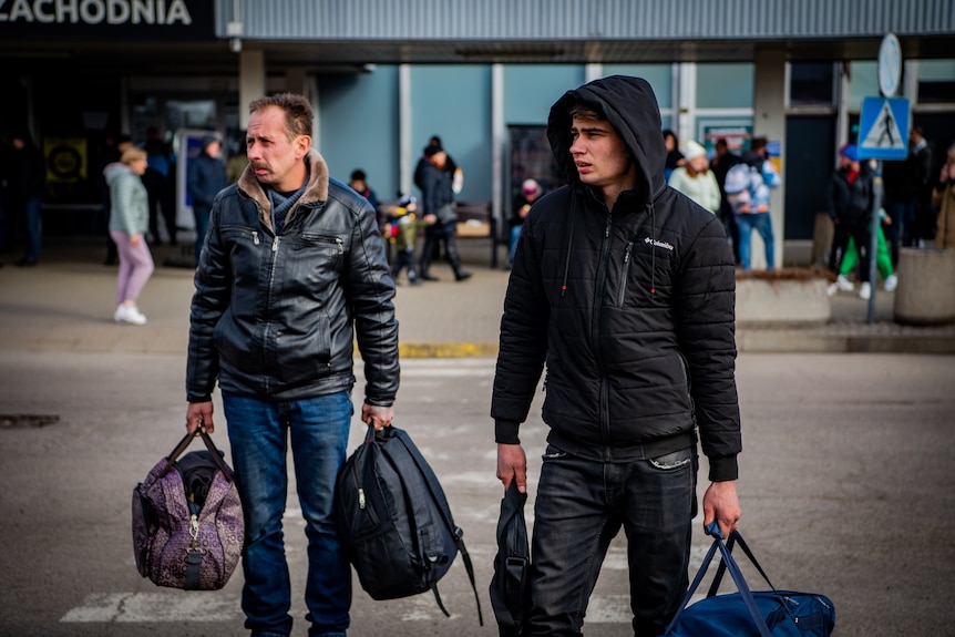 Two men stand on a road holding their bags as they wait for a bus.