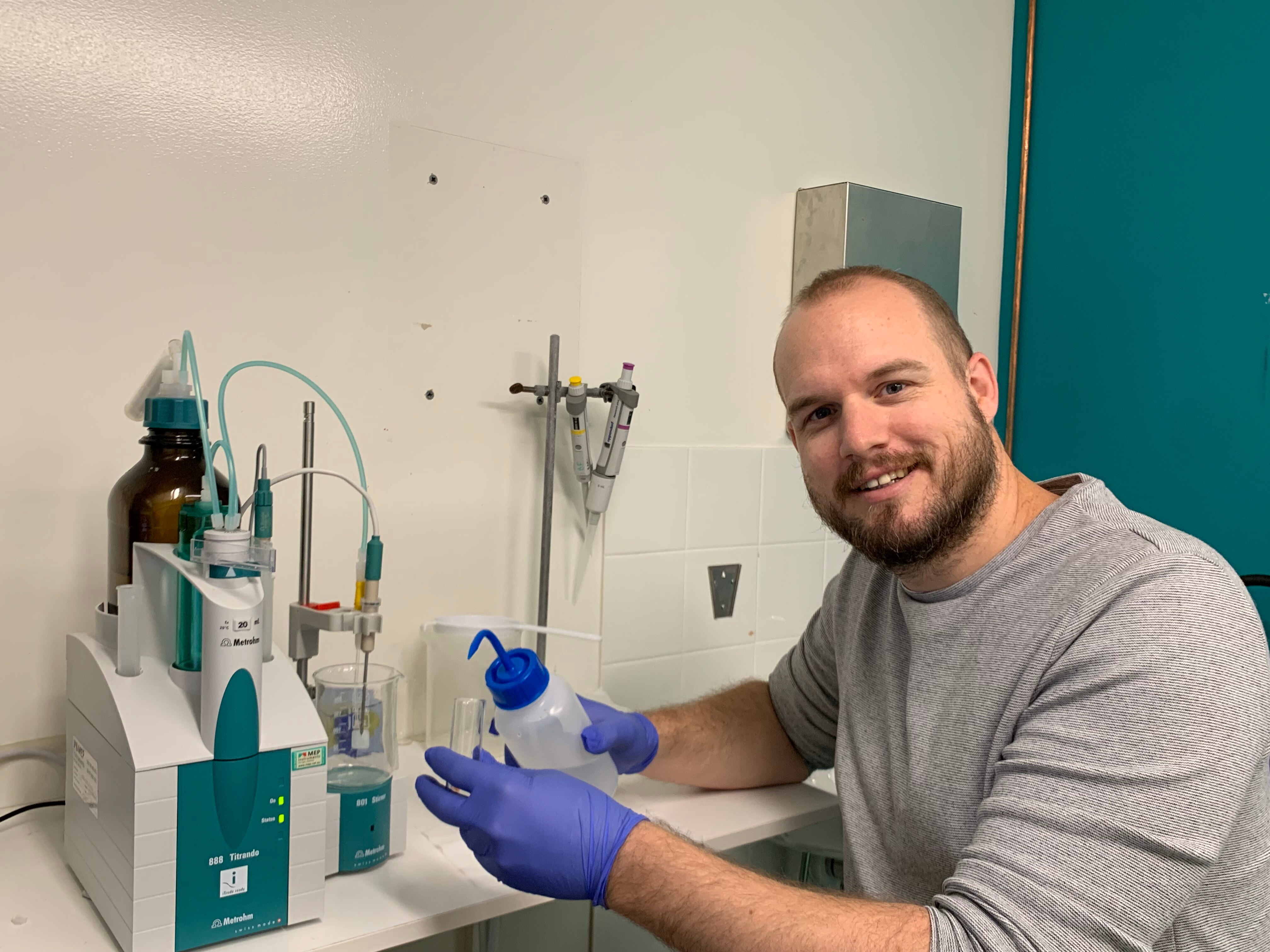 dr benjamin moss sits smiling in grey shirt in a science lab wearing gloves and pouring liquid into a beaker