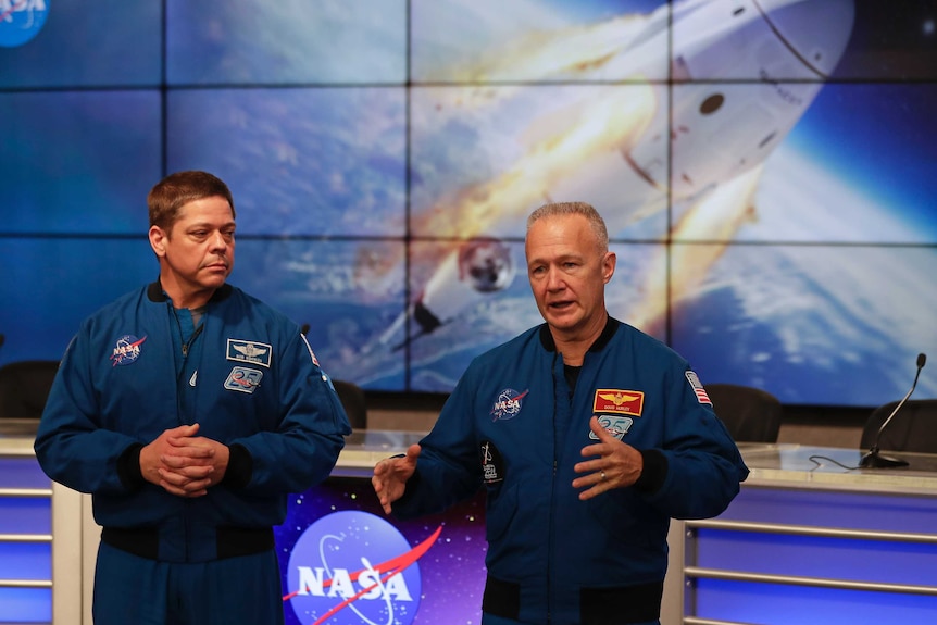 Two men in NASA uniforms speak in front of a large screen