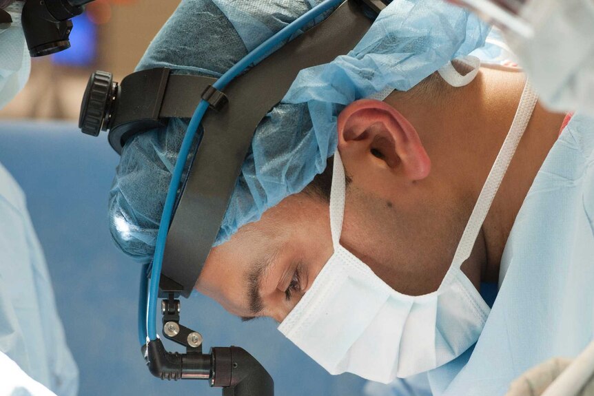 Doctor wearing mask, magnifying glass and cap leans over patient