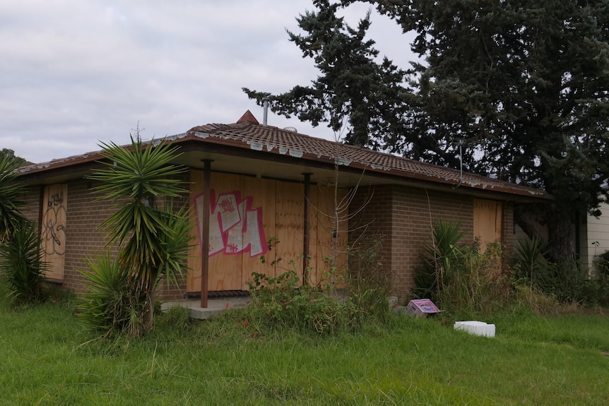 A low brick house with boarded up windows and rubbish in long grass