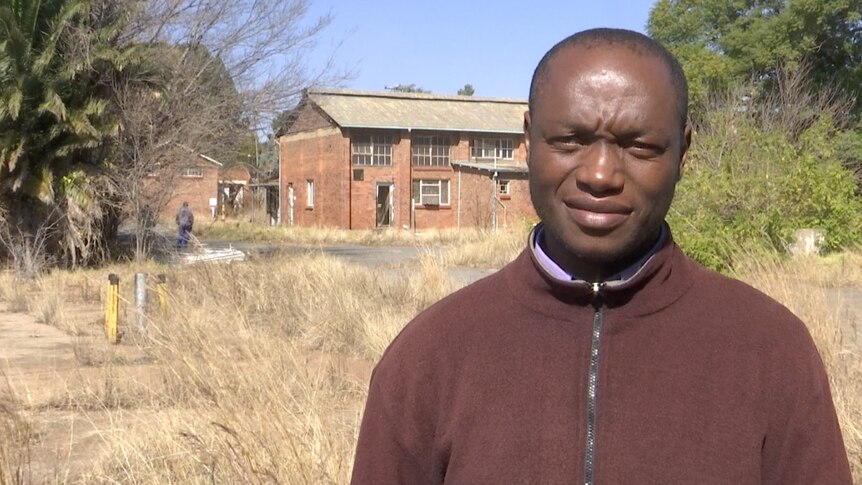 Comfort Zwane, wearing a brown zip-up jumper, stands before a dilapidated brick building