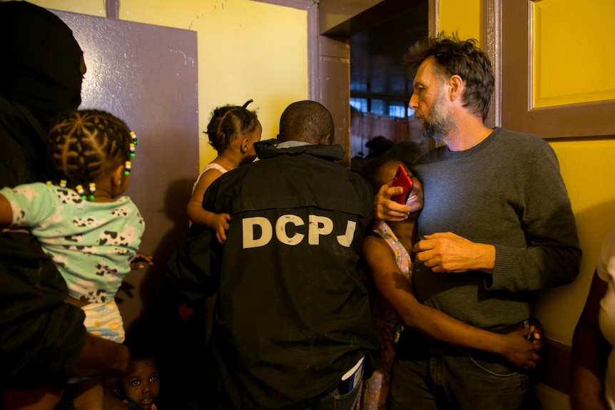 A police officer takes a child in his arms through a door as another child clings to a man in a pullover and jeans.