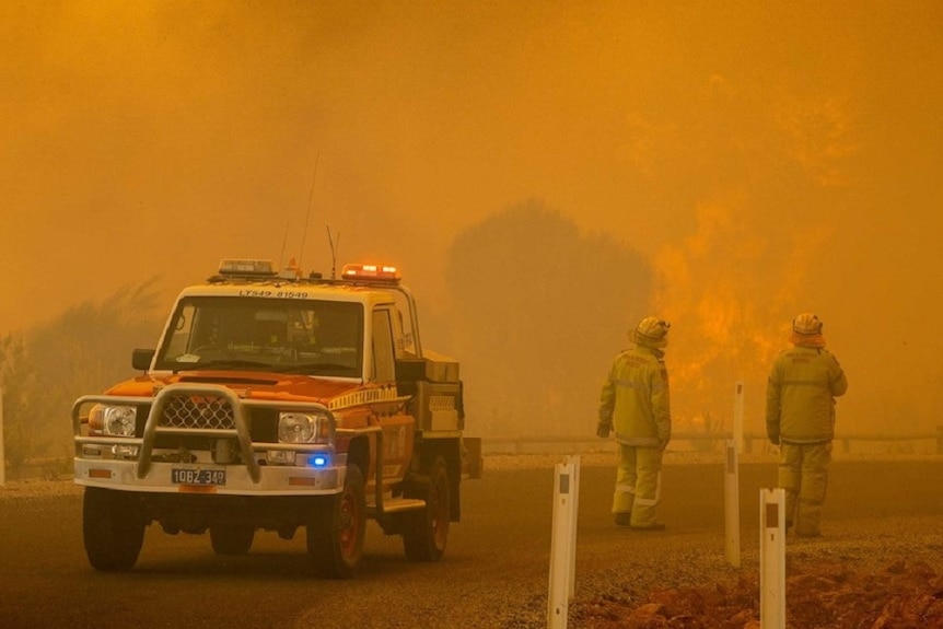 A small fire truck and 2 firefighters seen through an orange smoke haze.