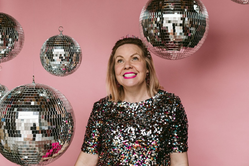 Kate Forsyth wears a sequin top, standing against a pink wall with disco balls above her.