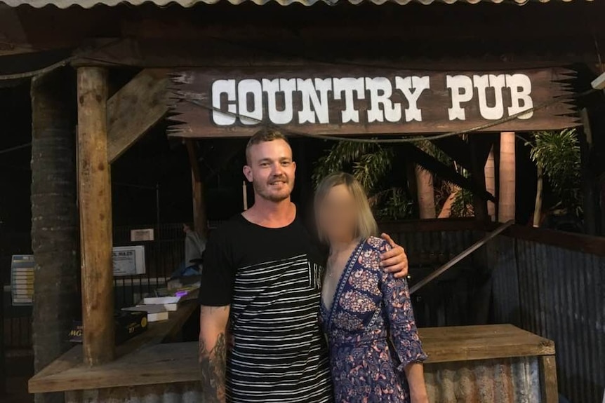 A man and woman stand together under a wooden sign with the words "Country pub".