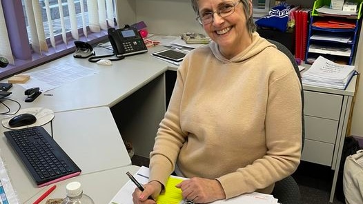 A woman in a yellow jumper and glasses smiles at a desk 