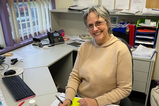 A woman in a yellow jumper and glasses smiles at a desk 