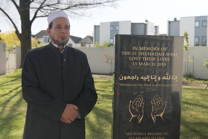 A man dressed in a suit stands with his hands clasped beside a plaque in Christchurch.