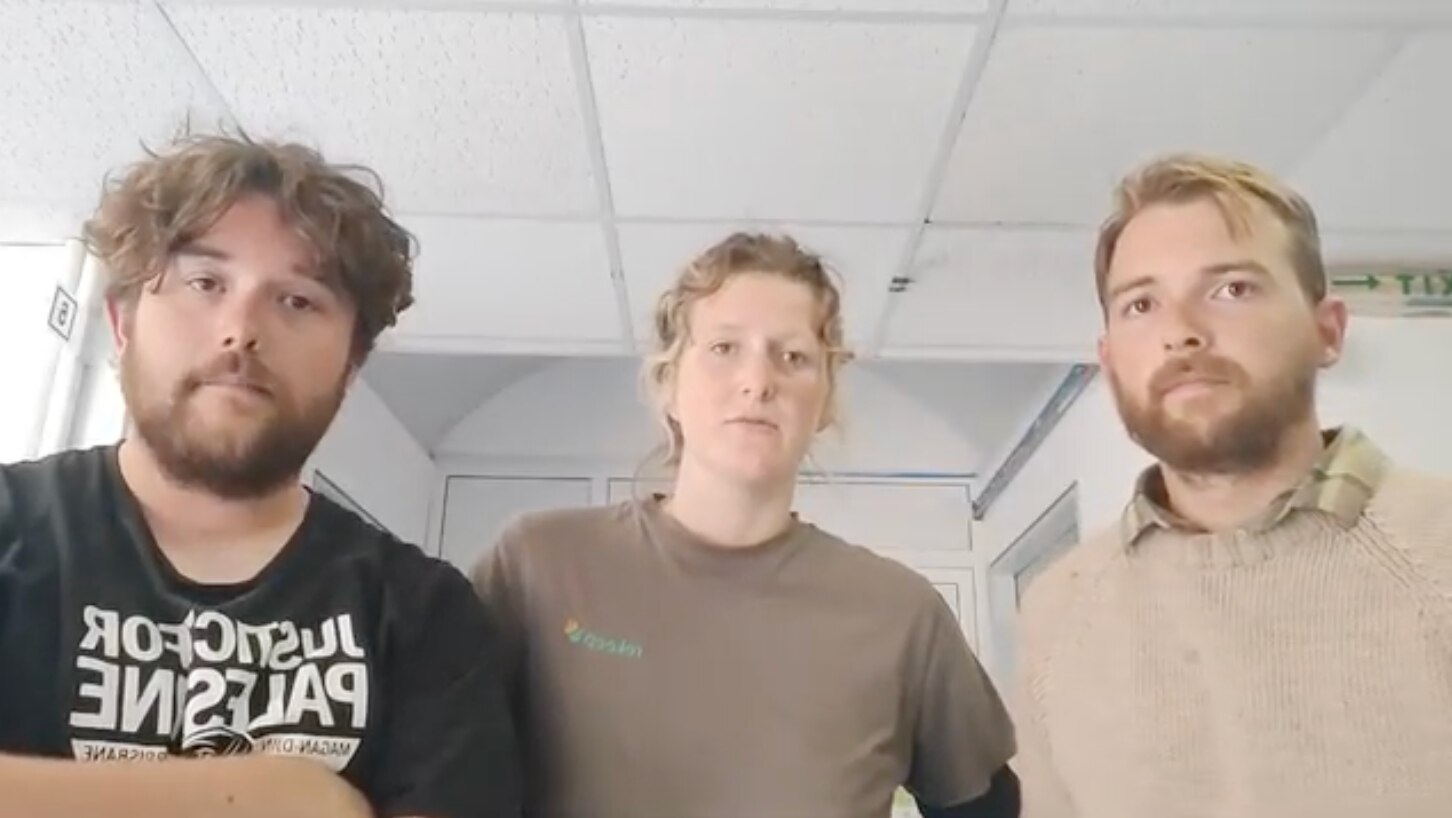 Two young men and a young woman look seriously at the camera inside a grey-walled room.