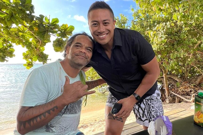 Two Pacific Island men standing on a beach in the shade smiling after receiving a tatau