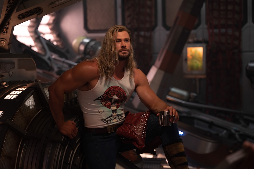 Ripped young white man with long blond hair in tight muscle tank sitting in arcade.