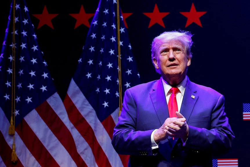 Donald Trump clasps his hands while bathed in purple lights 
