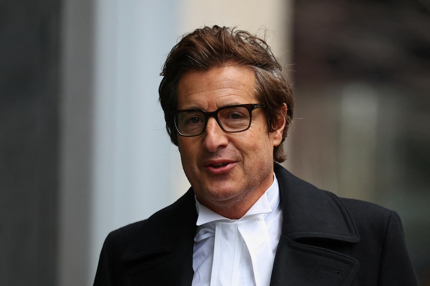 A man weraing glasses and a white shirt with a tight collar and black jacket
