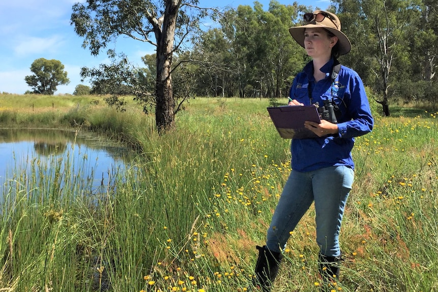 A woman standing next to a dam in a paddock writes on a clipboard. 