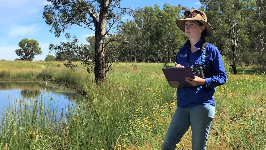 A woman standing next to a dam in a paddock writes on a clipboard. 