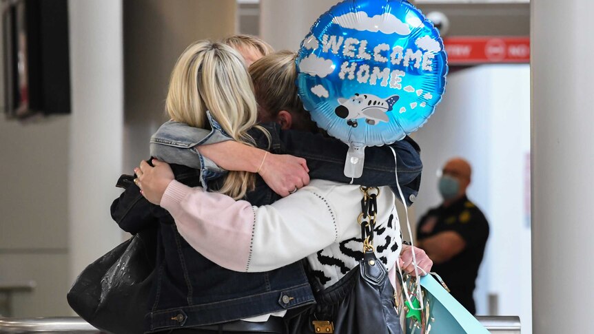 Three women embrace at the airport. One is calling a balloon that says "welcome home"