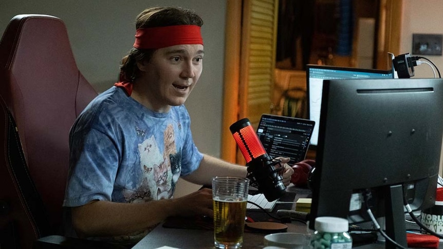 A man sitting in front of a computer, wearing a colourful T-shirt and a red head-band.