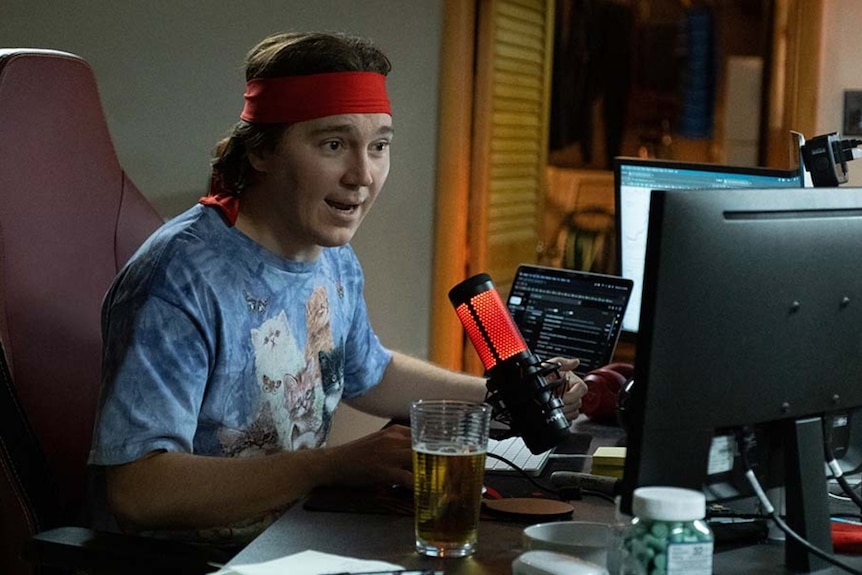 A man sitting in front of a computer, wearing a colourful T-shirt and a red head-band.