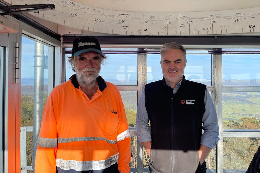 two men standing in a tower with a map behind them, one in high vis