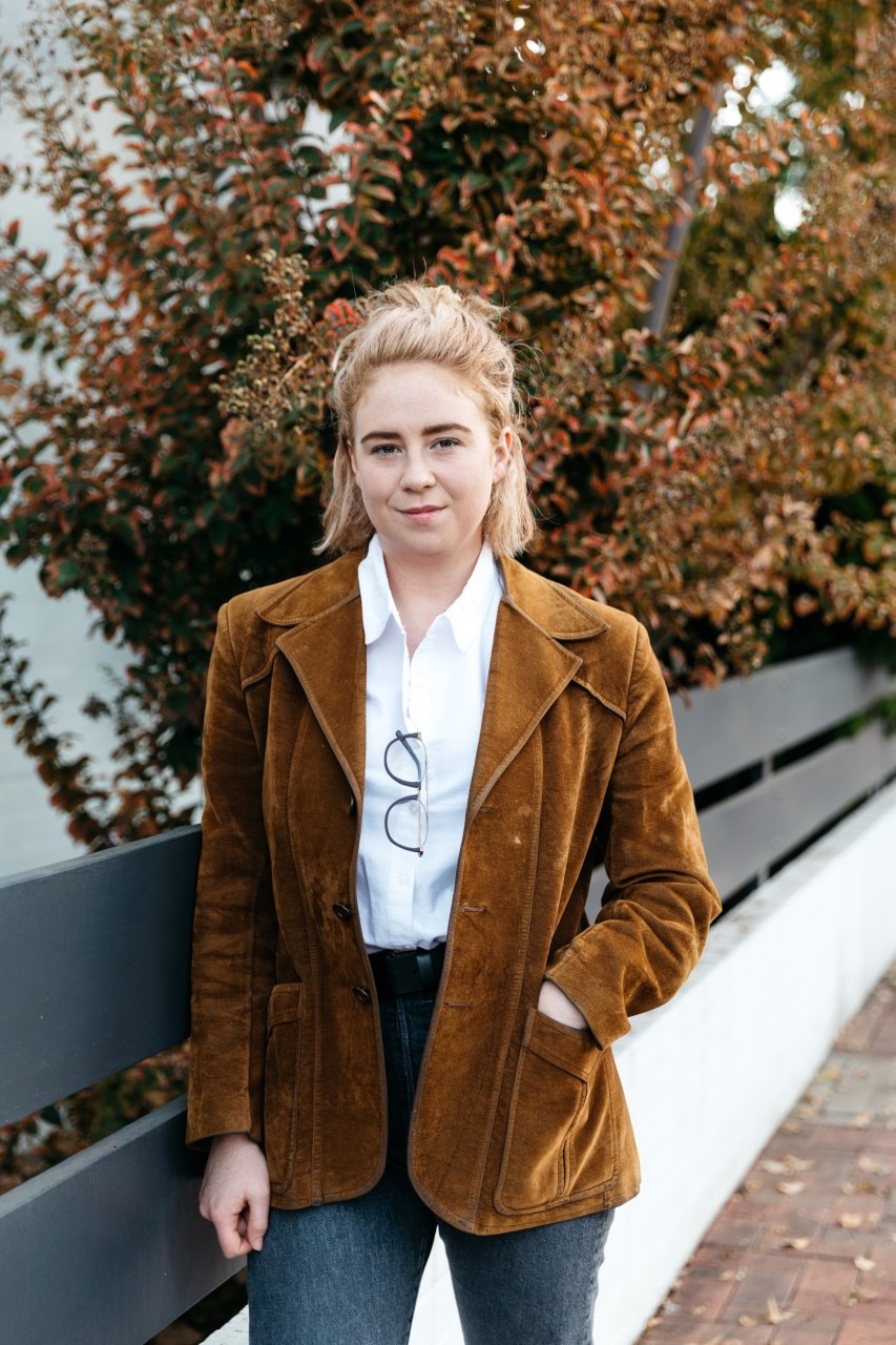Abbey Kendall stands in front of a park bench and a tree.