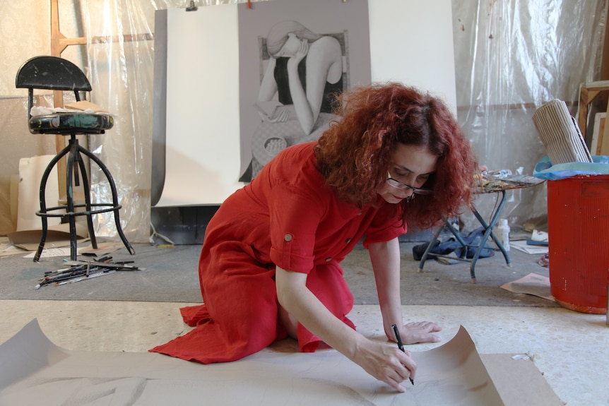 A woman in a red dress knees on the floor as she works on an artwork.