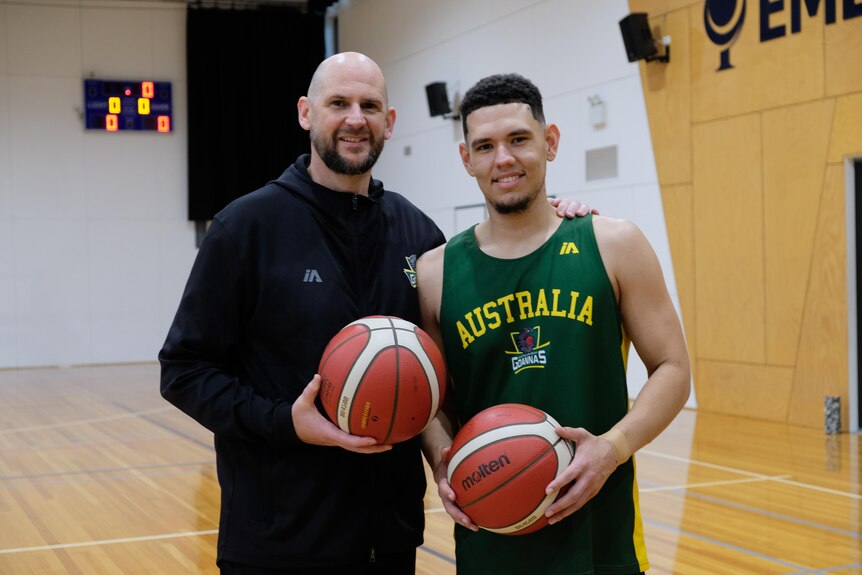 Brent and Jarrod hold basketballs and smile to the camera. Brent has his arm around Jarrod, a scoreboard is in the background.