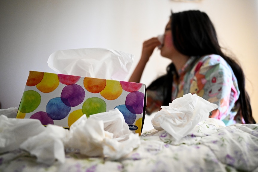 a young woman sick with a cold sitting on a bed blowing her nose with a tissue 