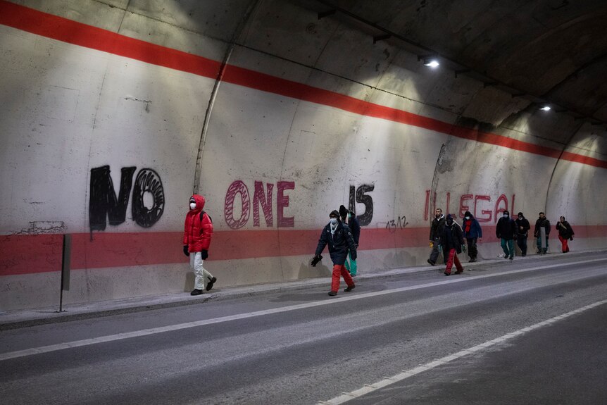 Migrants walk past graffiti that reads "No One is Illegal" in a tunnel.