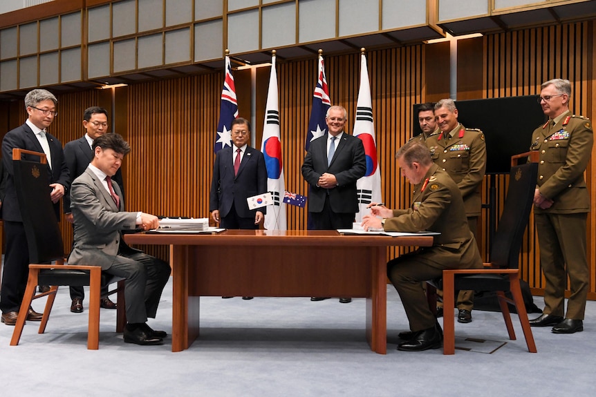 Two men sit at a table opposite each other surrounded by officials. 