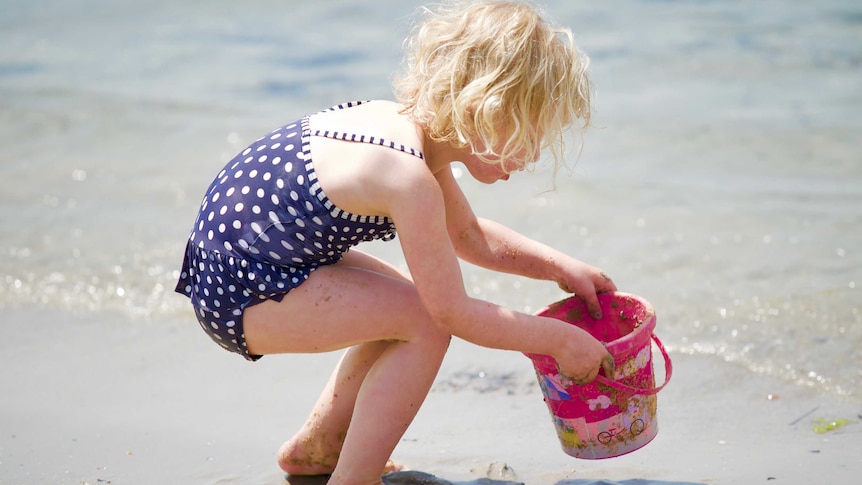 A young girl plays with a sand bucket at the edge of the water at Williamstown beach.