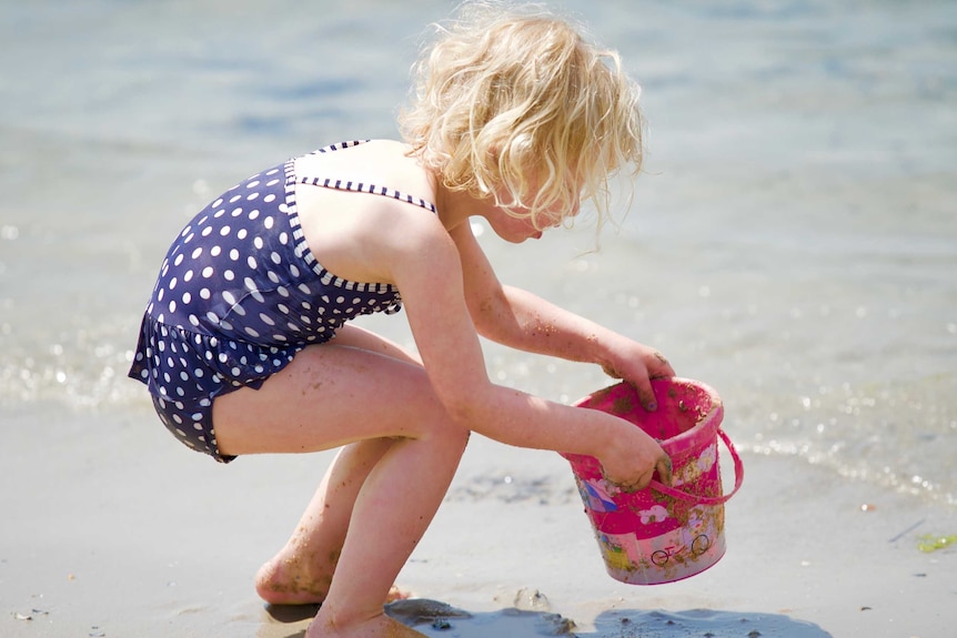 A young girl plays with a sand bucket at the edge of the water at Williamstown beach.