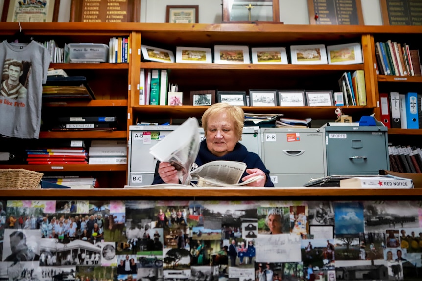 Older woman sits at desk covered in photos with cluttered bookshelf behind her