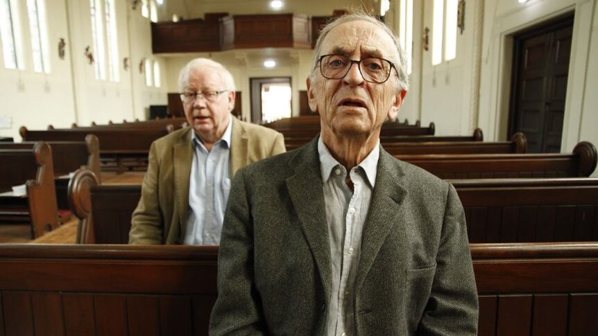 Two older men sit in church pews, dressed in suits.