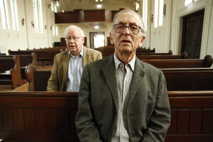 Two older men sit in church pews, dressed in suits.