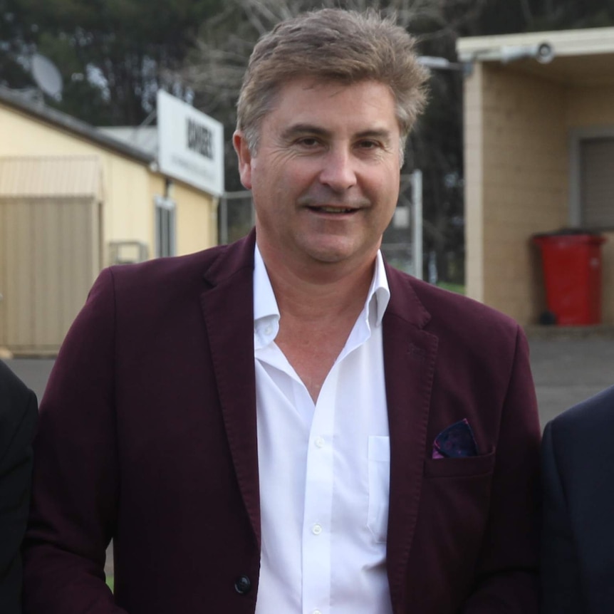 A man in a jacket and collared shirt smiles at the camera.