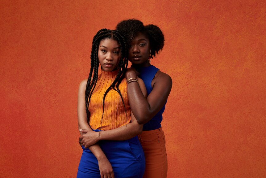 Two Black women stand in front of an orange background. One stands behind the other, leaning against her shoulder. 