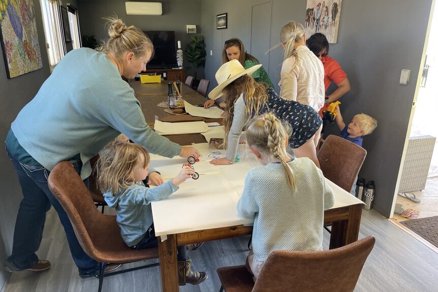 Children and parents busily lean over a table with drawing materials on it.