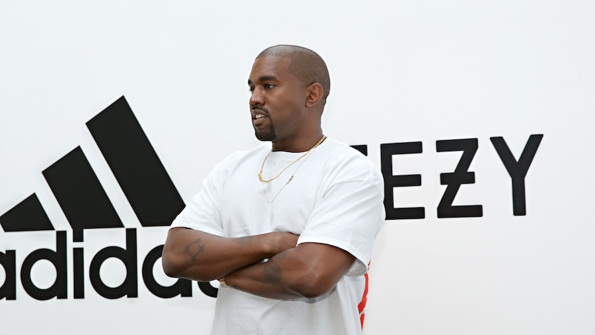 Ye wearing white and standing with arms crossed in front of an adidas and Yeezy sign